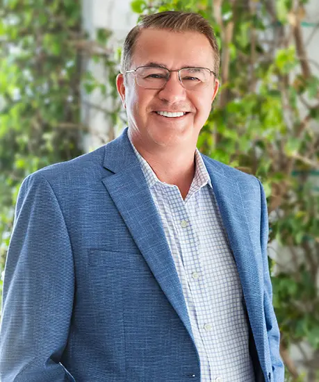 Don Rothrock is smiling in front of an ivy covered wall wearing a blue sports jacket and a blue and white checkered button up.